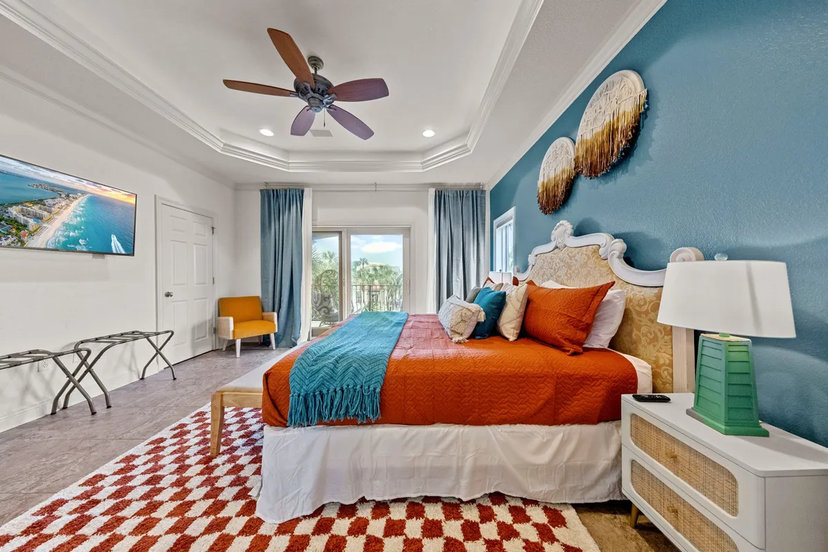 A spacious master bedroom featuring a large bed adorned with colorful, inviting bedding. Sunlight pours in through wide windows offering an ocean view, while tasteful artwork and a textured area rug contribute to a serene, comfortable atmosphere.
