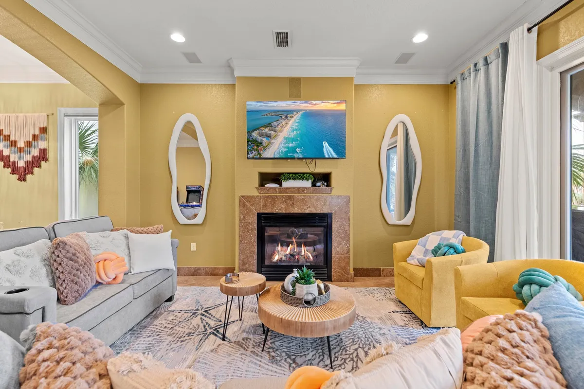 A cozy living area anchored by a fireplace with a mantel and TV above, flanked by two unique mirrors. Comfortable seating includes a grey sofa and bright yellow armchairs set atop a patterned area rug, ideal for socializing or relaxing with family and friends.