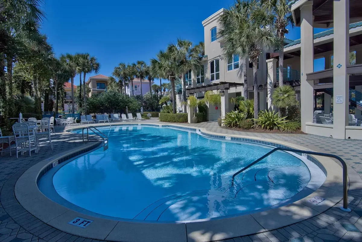 A large, heated swimming pool surrounded by swaying palm trees and poolside chairs, with an adjacent building offering shaded patio seating. The setting is inviting and family-friendly, perfect for swimming, lounging, and socializing in a resort-style environment.