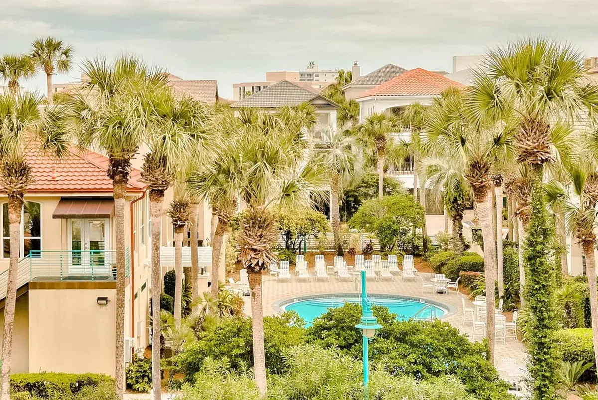 The image showcases a sparkling heated pool surrounded by inviting lounge chairs, lush palm trees, and meticulously landscaped grounds. Mediterranean-style homes add to the upscale, tropical atmosphere, while the tranquil setting suggests a private, resort-like escape just steps from the beach.