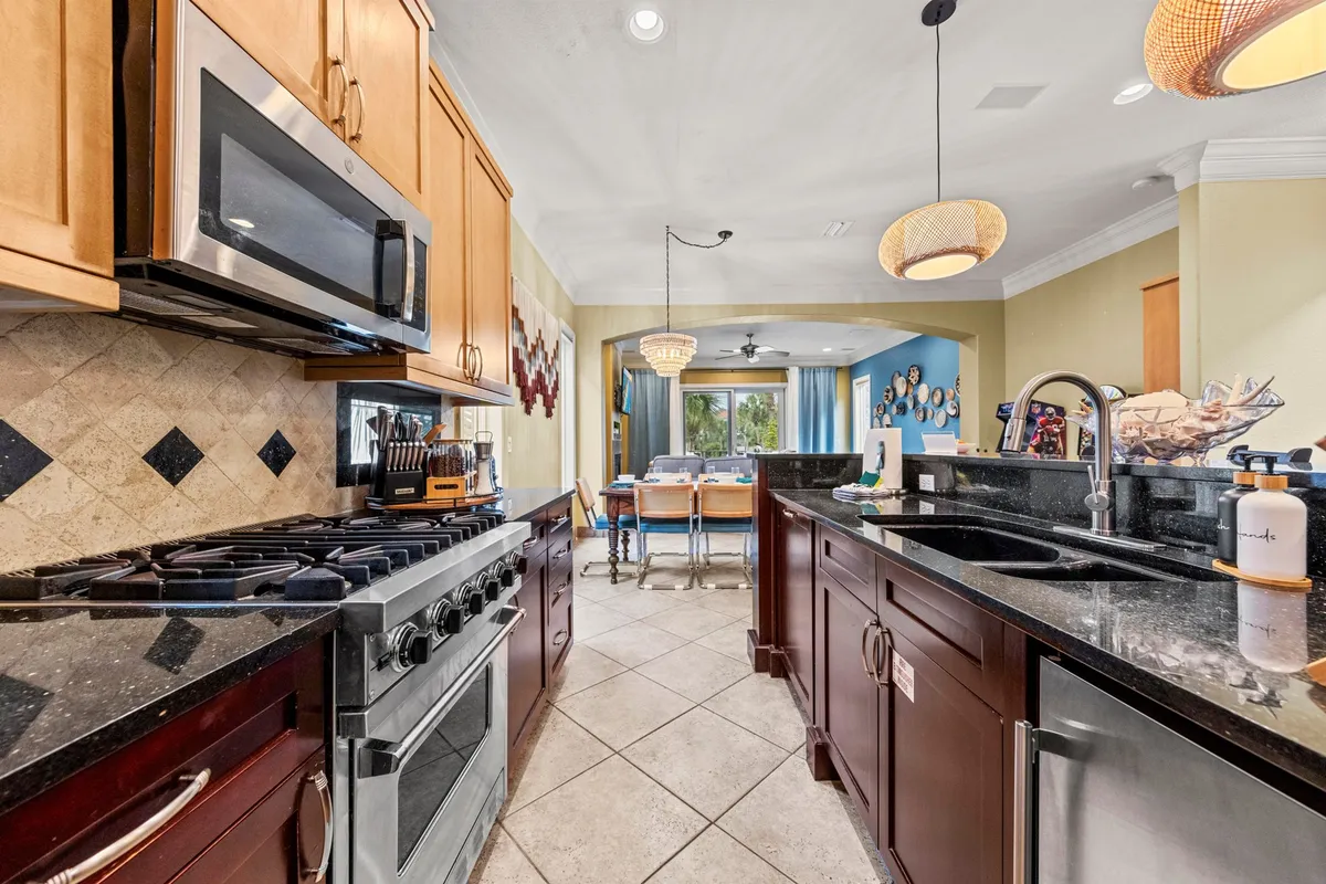 A spacious kitchen featuring a modern stainless steel stove and oven, dark granite kitchen countertops, and warm wooden cabinetry. The kitchen opens into a dining area with large windows and a blue accent wall, allowing natural light to fill the space.