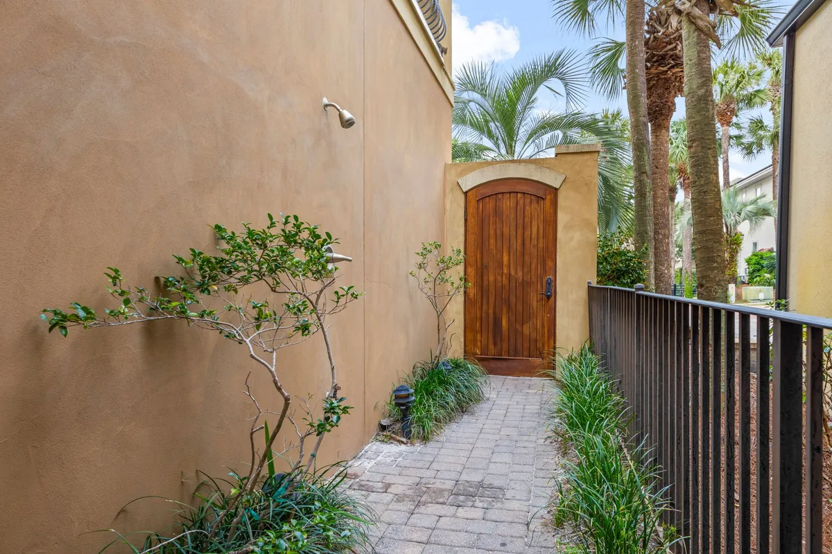 This exterior shot highlights a charming arched wooden door with an outdoor shower fixture nearby, set along a narrow brick path bordered by lush tropical plants and palm trees. The view evokes a sense of entering a private, tranquil coastal haven.