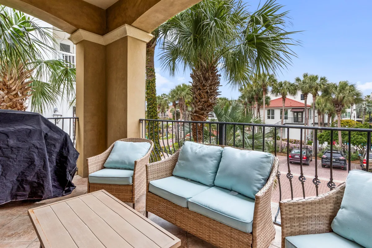 A covered balcony with arch design, equipped with wicker patio furniture featuring light blue cushions and a black grill with a protective cover. Lush palm trees surround the space, offering privacy and a refreshing tropical atmosphere for outdoor dining or lounging.