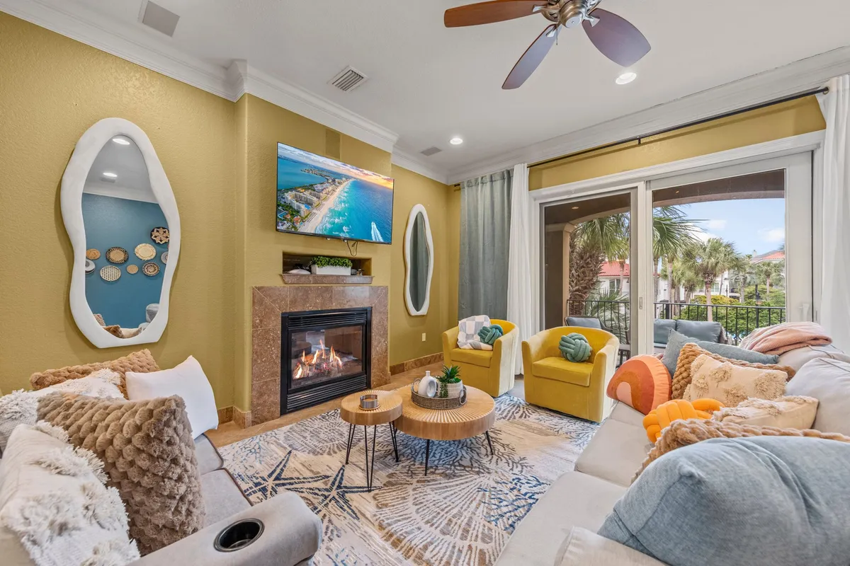 A spacious, sunlit living room featuring a gas fireplace with a mantle and TV above, a large sectional sofa with colorful cushions, and bright yellow armchairs. Sliding glass doors let in abundant natural light and offer a view of palm trees and a balcony, creating an inviting and lively communal space.