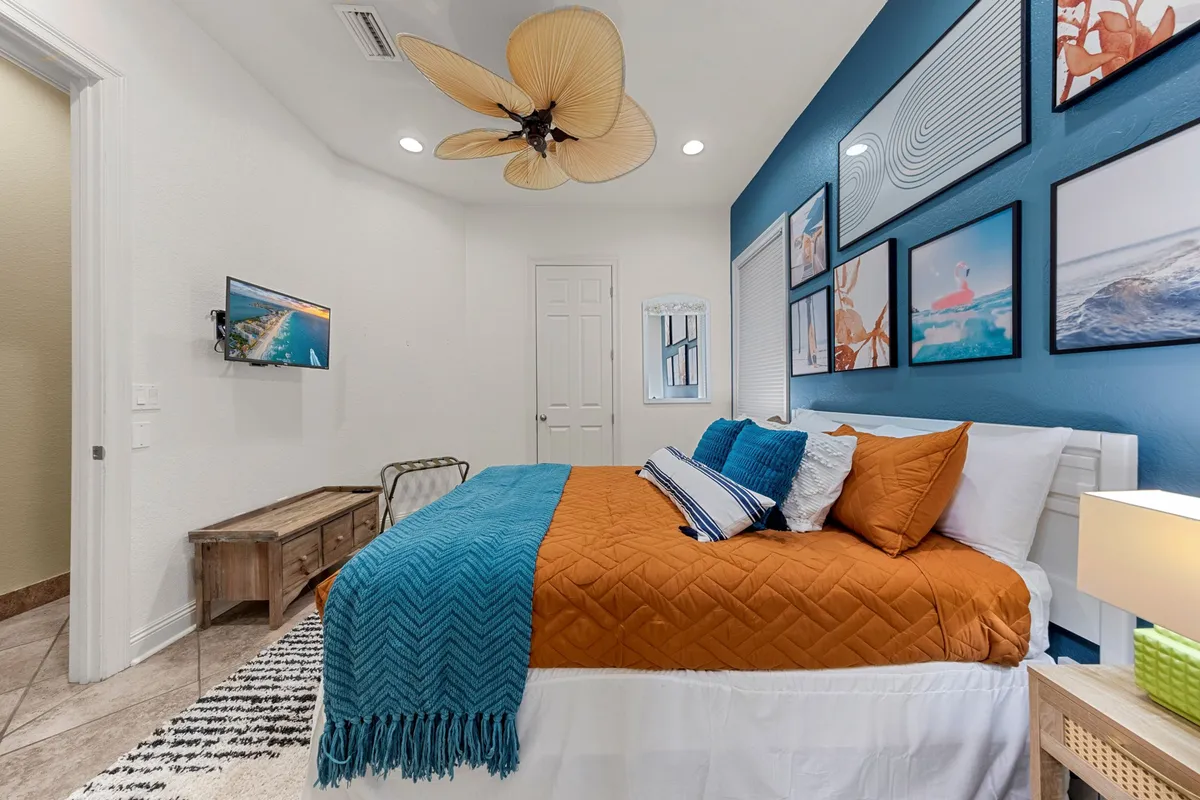 A masterfully appointed bedroom with a vibrant blue accent wall, ocean-themed artwork, an orange and blue bed, a unique ceiling fan with leaf-shaped blades, and a wall-mounted flat-screen TV. The room radiates calm and rejuvenation, offering both style and comfort.