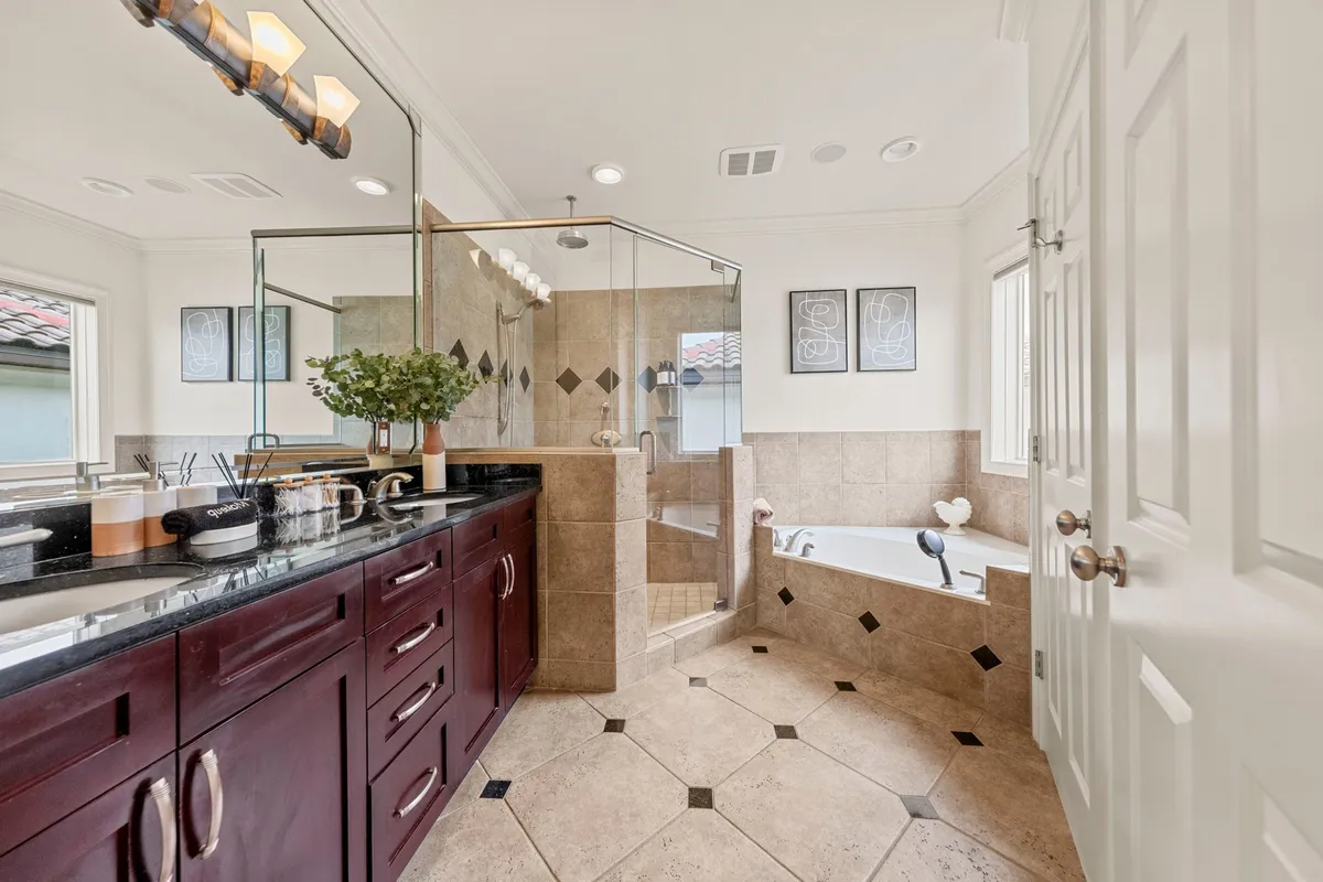A luxurious bathroom showcasing a deep soaking bathtub, a spacious glass-enclosed shower, and a double vanity with dark wood cabinetry and granite countertop. Bright natural light pours in from a window, illuminating the spa-like finishes and serene atmosphere.