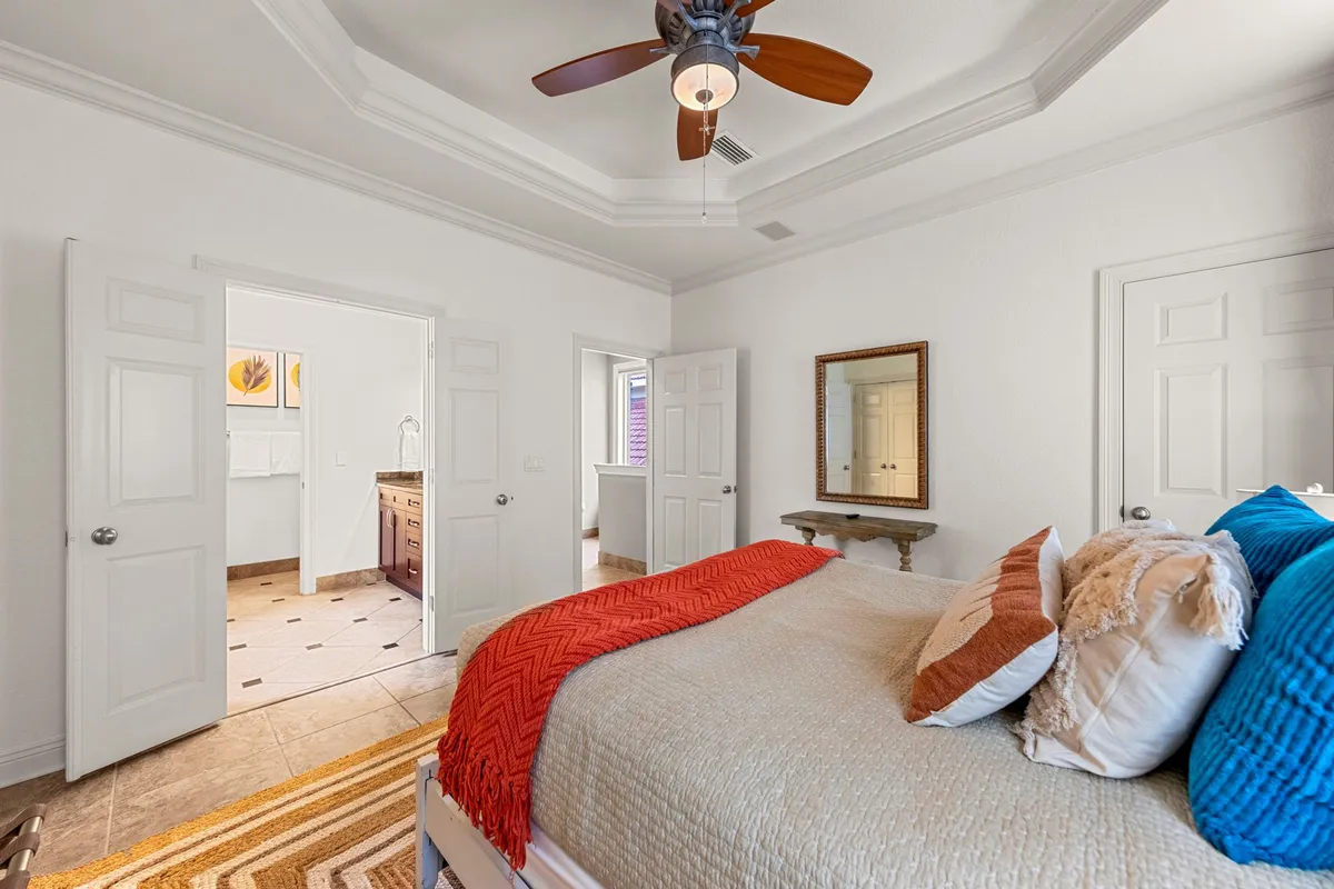 A spacious bedroom featuring a large bed adorned with colorful pillows and a throw blanket. A ceiling fan with wooden blades hangs overhead, and open double doors lead directly to a sleek bathroom. The room feels tranquil and cozy, with a framed mirror above a console table and soft, natural light enhancing the restful ambiance.