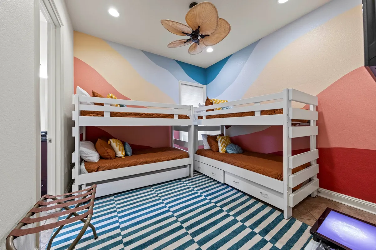 A cheerful and thoughtfully designed bedroom featuring white wooden bunk beds with brown bedding and colorful pillows. The space includes a striped blue and white rug, a geometric pastel wall mural, and a ceiling fan with leaf-shaped blades, creating a playful and welcoming environment for children or extra guests.