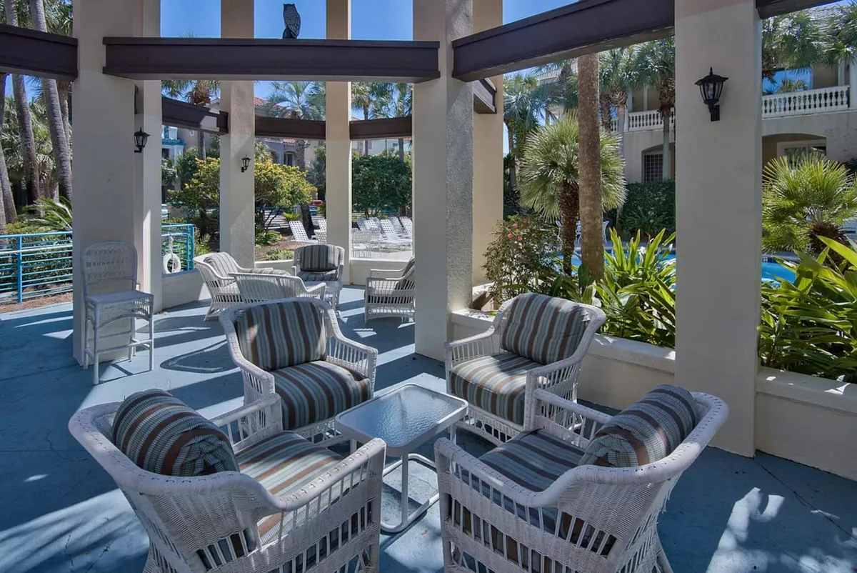 A serene outdoor seating area featuring white wicker chairs with striped cushions, a glass-topped table, and tall pillars. Lush tropical plants and palm trees surround the space, creating a peaceful setting for casual conversation, games, or simply enjoying the garden view.