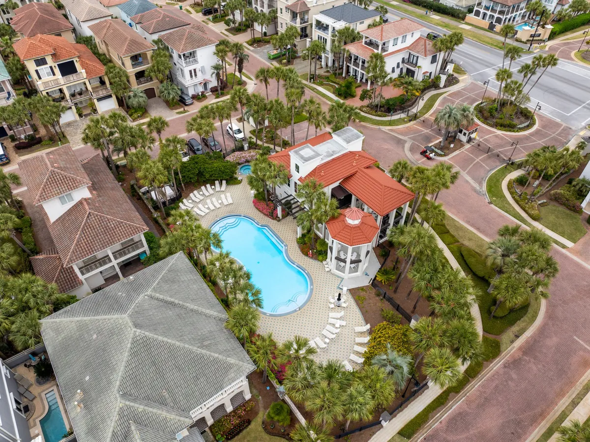A beautifully landscaped heated pool and spa area, complete with poolside lounge chairs and surrounded by tall palm trees. The inviting clubhouse with a red-tiled roof sits nearby, and the scene is bathed in tranquil sunlight, creating a welcoming space for all ages to relax and socialize.