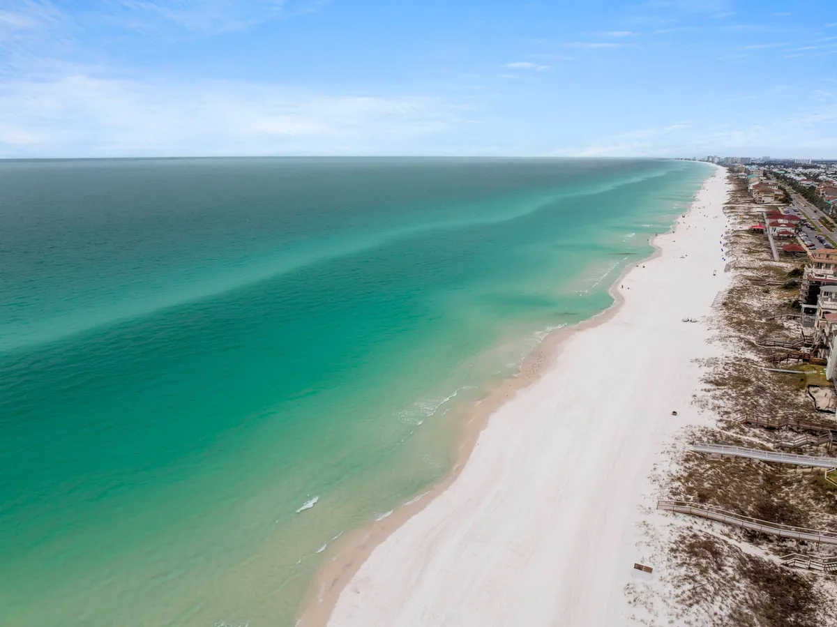 A stunning stretch of pristine white sandy beach with turquoise ocean waters, set against a backdrop of distant beachfront properties and a clear blue sky. The beach is uncrowded, emphasizing exclusivity and privacy for residents and guests.
