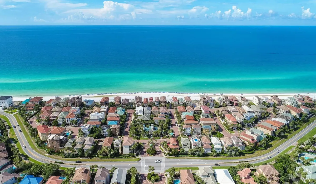 An aerial view of the private beach with turquoise waters, rows of residential homes, a main road leading into the secure community, and lush landscaping. The image highlights the peaceful, less-crowded beachfront and the inviting neighborhood atmosphere.
